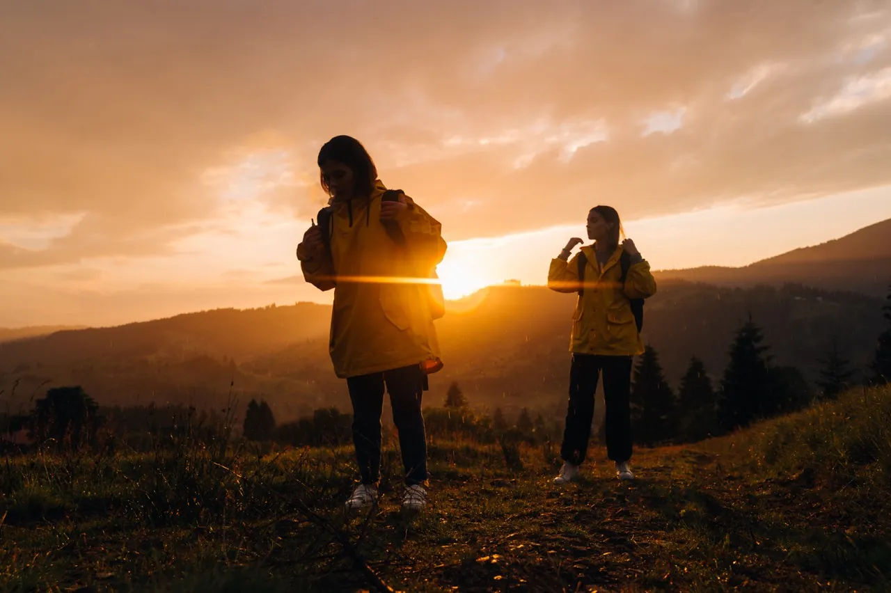 Two girls hikers in yellow jackets climb the mountain in. rain on a background of beautiful sunset. Two tourists in raincoats with backpacks go hiking at sunset.